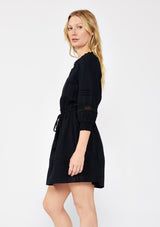 [Color: Black] A blonde model wearing a ultra fall bohemian mini dress in black. With a button front, long sleeves with smocked cuffs, tassel tie waist detail, and lace inserts.