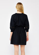 [Color: Black] A blonde model wearing a ultra fall bohemian mini dress in black. With a button front, long sleeves with smocked cuffs, tassel tie waist detail, and lace inserts.