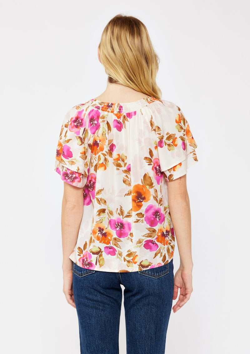 [Color: Natural/Orchid] A blonde model wearing a relaxed fit top in a floral print. With short double flutter sleeves, split v neckline, and a smocked detail at the neck. A dressy but casual floral top styled with dark denim.