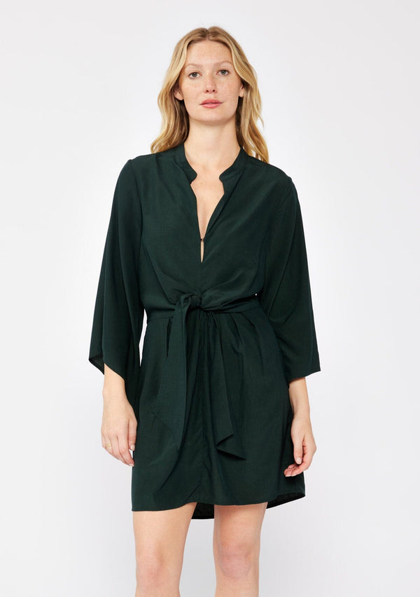 [Color: Olive] A blonde model wearing a green dark olive mini dress. With a deep v-neckline, front hook and eye closure, front tie waist detail, flared long sleeves , and a relaxed fit. A dressy but casual fall dress. 