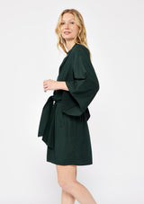 [Color: Olive] A blonde model wearing a green dark olive mini dress. With a deep v-neckline, front hook and eye closure, front tie waist detail, flared long sleeves , and a relaxed fit. A dressy but casual fall dress.