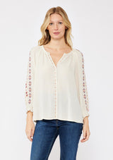 [Color: Natural/Coral/Plum] A blonde model wearing a beige ivory bohemian blouse with embroidered details. A relaxed fit blouse with a split v neckline, long sleeve, and shirred wrist cuffs. Styled with dark denim jeans for a laid back fall look. 