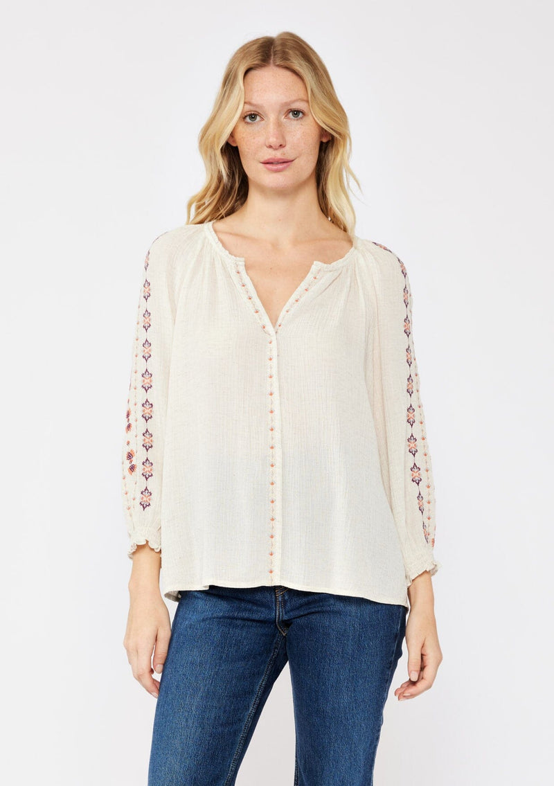 [Color: Natural/Coral/Plum] A blonde model wearing a beige ivory bohemian blouse with embroidered details. A relaxed fit blouse with a split v neckline, long sleeve, and shirred wrist cuffs. Styled with dark denim jeans for a laid back fall look. 