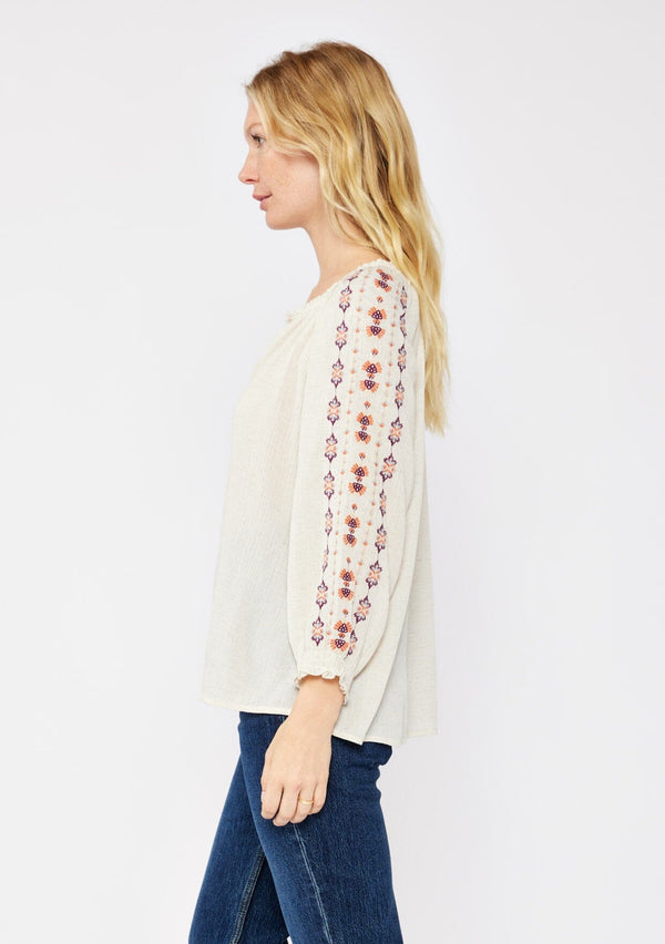 [Color: Natural/Coral/Plum] A blonde model wearing a beige ivory bohemian blouse with embroidered details. A relaxed fit blouse with a split v neckline, long sleeve, and shirred wrist cuffs. Styled with dark denim jeans for a laid back fall look.