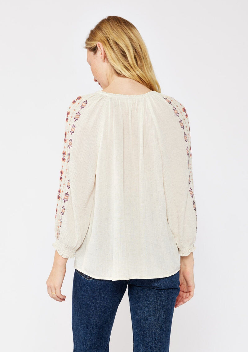 [Color: Natural/Coral/Plum] A blonde model wearing a beige ivory bohemian blouse with embroidered details. A relaxed fit blouse with a split v neckline, long sleeve, and shirred wrist cuffs. Styled with dark denim jeans for a laid back fall look.