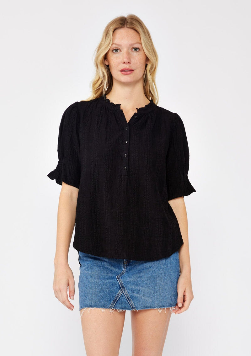 [Color: Black] A front facing image of a blonde model wearing a black bohemian cotton blend blouse in a textured gingham. With short puff sleeves, a button front, a high ruffled neckline, and smocked elastic details at the sleeve.