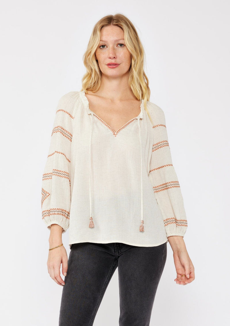 [Color: Natural/Rust] A blonde model wearing a beige ivory blouse with bohemian embroidered details. A relaxed fit blouse with a split v neckline and tassel tie front, long sleeves, and ruffle trim details at the neck. Styled with dark denim for an ultra boho look for the fall season. 