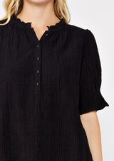 [Color: Black] A detail image of a blonde model wearing a black bohemian cotton blend blouse in a textured gingham. With short puff sleeves, a button front, a high ruffled neckline, and smocked elastic details at the sleeve.