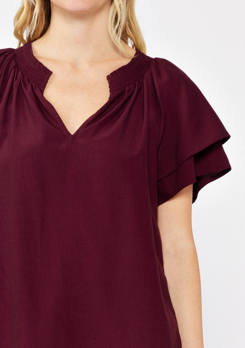 [Color: Merlot] A blonde model wearing a sophisticated burgundy wine top. With double short flutter sleeves, smocked neck detail, split v neck, and a relaxed fit. An everyday top perfect for the office or a casual outing.