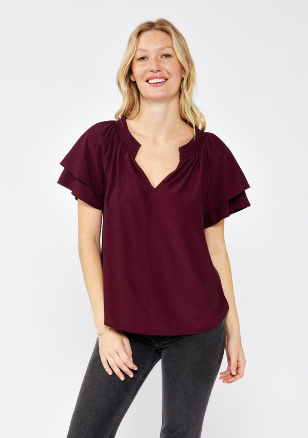 [Color: Merlot] A blonde model wearing a sophisticated burgundy wine top. With double short flutter sleeves, smocked neck detail, split v neck, and a relaxed fit. An everyday top perfect for the office or a casual outing. 