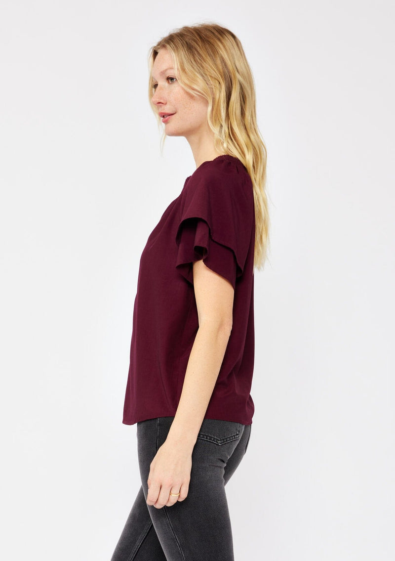 [Color: Merlot] A blonde model wearing a sophisticated burgundy wine top. With double short flutter sleeves, smocked neck detail, split v neck, and a relaxed fit. An everyday top perfect for the office or a casual outing.