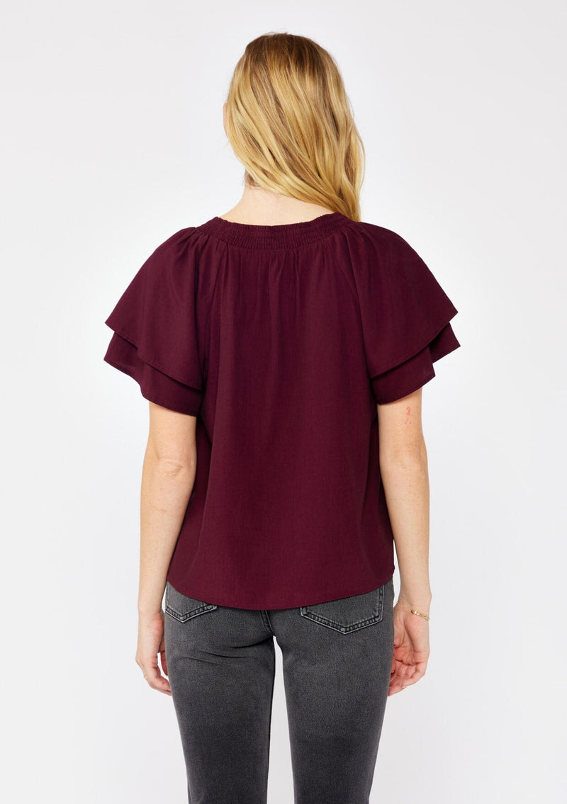 [Color: Merlot] A blonde model wearing a sophisticated burgundy wine top. With double short flutter sleeves, smocked neck detail, split v neck, and a relaxed fit. An everyday top perfect for the office or a casual outing.