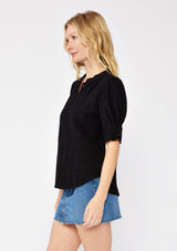 [Color: Black] A side facing image of a blonde model wearing a black bohemian cotton blend blouse in a textured gingham. With short puff sleeves, a button front, a high ruffled neckline, and smocked elastic details at the sleeve.