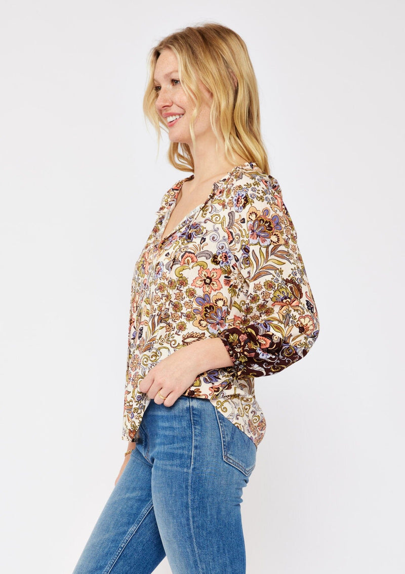 [Color: Natural/Brown] A blonde model wearing a neutral brown top with mixed florals for the fall season. With a split v neckline and tie neck, long sleeve, elastic cuff, and a relaxed fit. The perfect fall top paired with denim jeans.