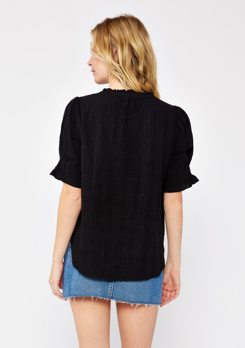 [Color: Black] A back facing image of a blonde model wearing a black bohemian cotton blend blouse in a textured gingham. With short puff sleeves, a button front, a high ruffled neckline, and smocked elastic details at the sleeve.