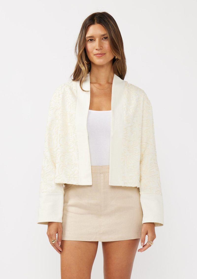 [Color: Vanilla] A front facing image of a brunette model wearing an off white cotton jacket with a soutache detailing. Featuring a collarless design, long sleeves, and an open front with a hook and eye closure. 