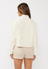 [Color: Vanilla] A back facing image of a brunette model wearing an off white cotton jacket with a soutache detailing. Featuring a collarless design, long sleeves, and an open front with a hook and eye closure. 