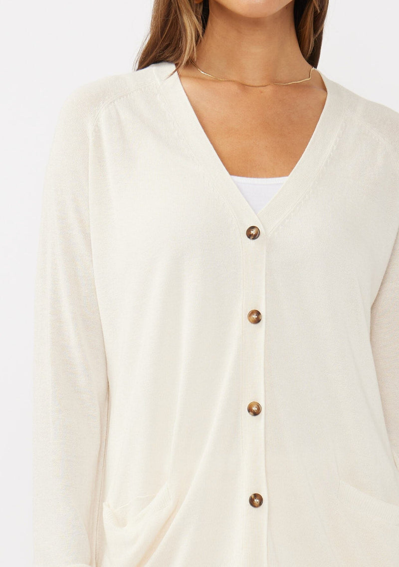 [Color: Vanilla] A detail image of a brunette model wearing an off white thin linen and viscose blend cardigan with long sleeves, a button front, and a v-neckline. An effortless basic throw on cardigan for the spring season. 