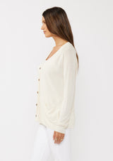 [Color: Vanilla] A side facing image of a brunette model wearing an off white thin linen and viscose blend cardigan with long sleeves, a button front, and a v-neckline. An effortless basic throw on cardigan for the spring season. 