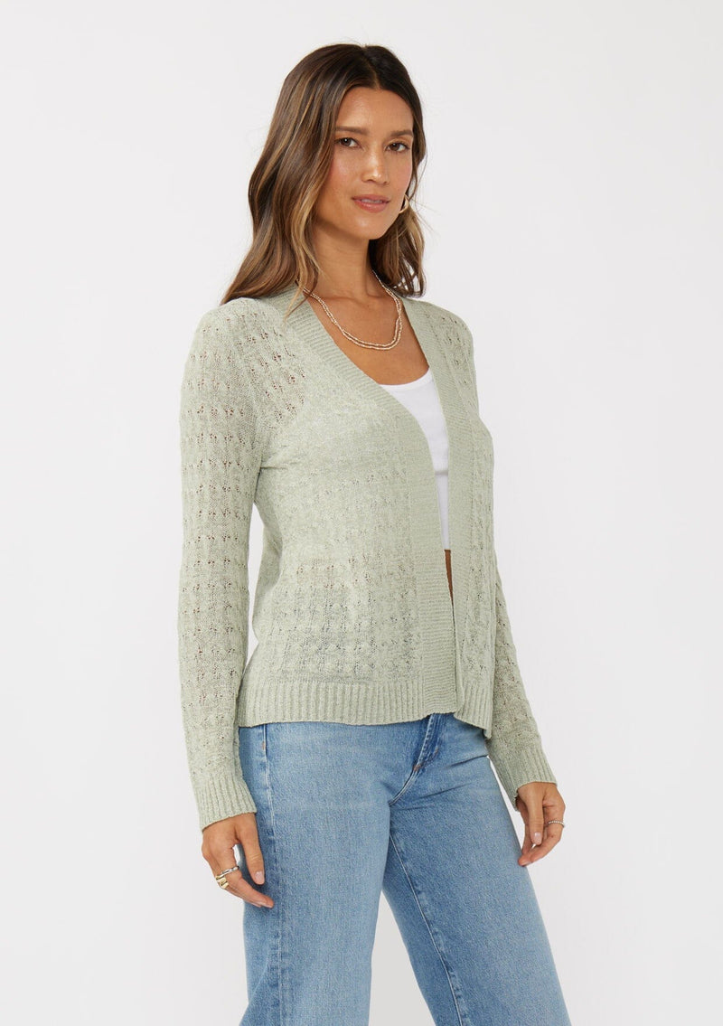 [Color: Light Sage] A side facing image of a brunette model wearing green tape yarn cardigan with open knit details. This lightweight cardigan features long sleeves, an open front, and ribbed trim, making it ideal for light layering in the spring.