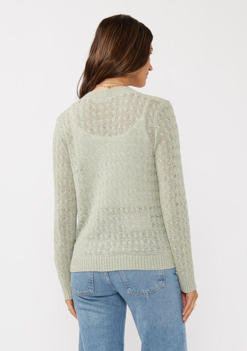 [Color: Light Sage] A back facing image of a brunette model wearing green tape yarn cardigan with open knit details. This lightweight cardigan features long sleeves, an open front, and ribbed trim, making it ideal for light layering in the spring.