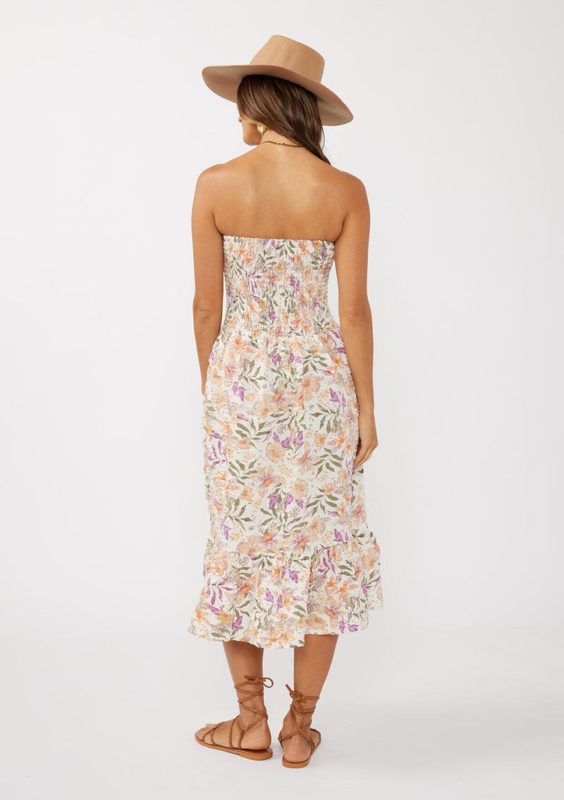 [Color: Cream/Dusty Lilac] A back facing image of a brunette model wearing a convertible floral midi dress with a puckered texture, smocked bodice, and ruffle trimmed hem. 