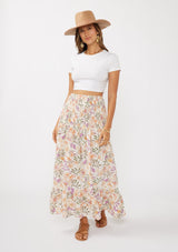 [Color: Cream/Dusty Lilac] A front facing image of a brunette model wearing a convertible floral maxi skirt with a puckered texture, smocked waist, and ruffle trimmed hem. 