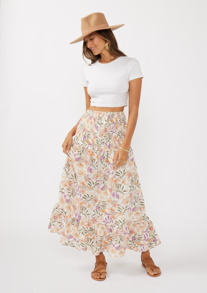 [Color: Cream/Dusty Lilac] A front facing image of a brunette model wearing a convertible floral maxi skirt with a puckered texture, smocked waist, and ruffle trimmed hem. 