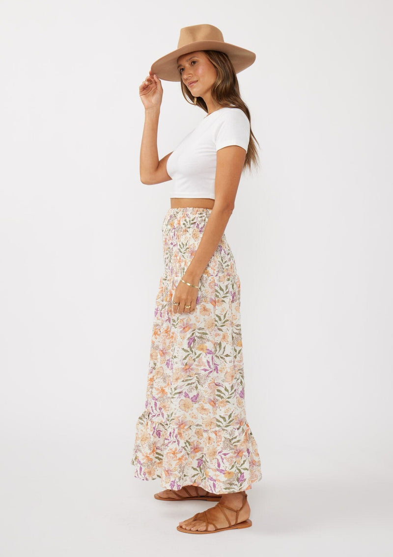 [Color: Cream/Dusty Lilac] A side facing image of a brunette model wearing a convertible floral maxi skirt with a puckered texture, smocked waist, and ruffle trimmed hem. 