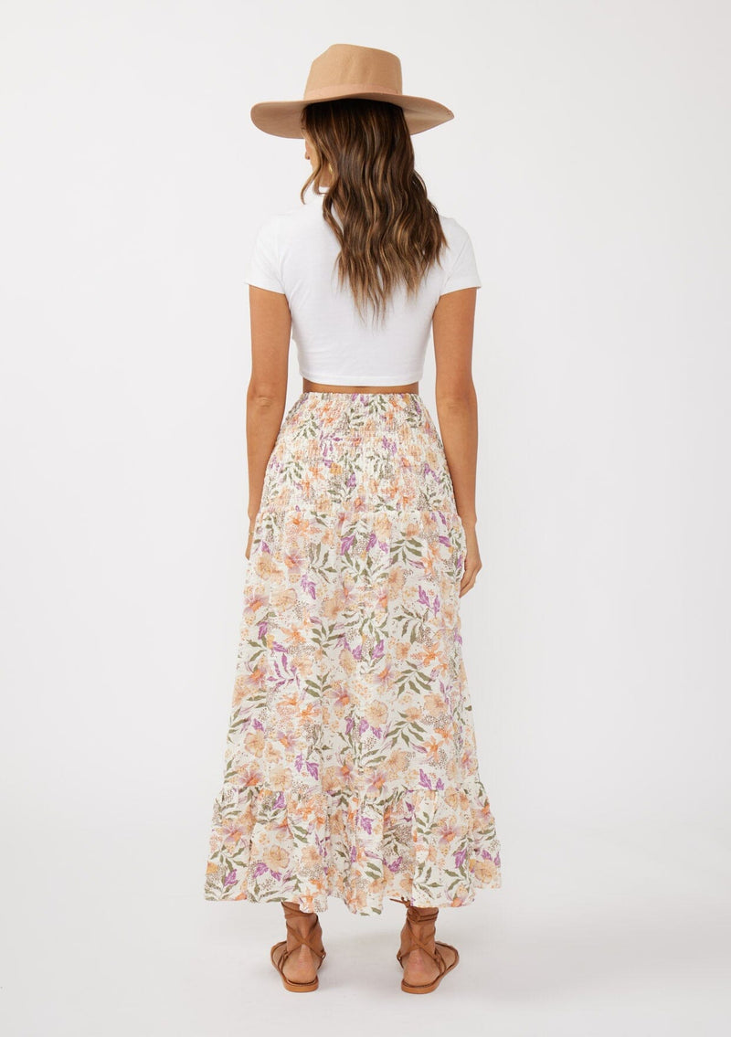 [Color: Cream/Dusty Lilac] A back facing image of a brunette model wearing a convertible floral maxi skirt with a puckered texture, smocked waist, and ruffle trimmed hem. 