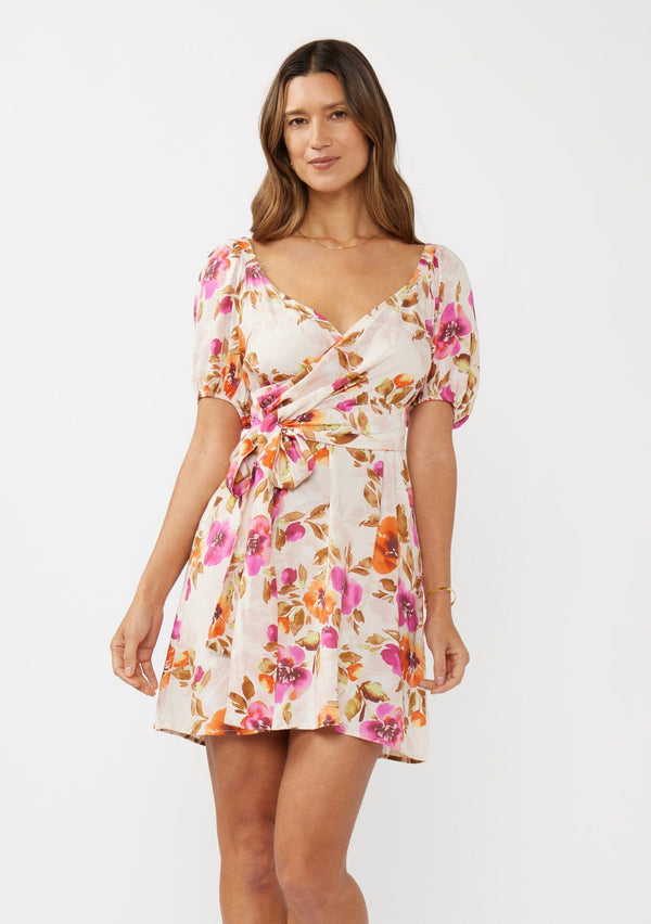 [Color: Natural/Orchid] A front facing image of a brunette model wearing a pretty floral mini dress in pink and orange. With a surplice v neckline, puff sleeves, attached waist belt with multiple styling opportunities, and smocked back. A cute floral dress for special occasions and dinner parties.