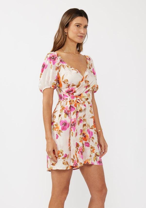 [Color: Natural/Orchid] A side facing image of a brunette model wearing a pretty floral mini dress in pink and orange. With a surplice v neckline, puff sleeves, attached waist belt with multiple styling opportunities, and smocked back. A cute floral dress for special occasions and dinner parties.