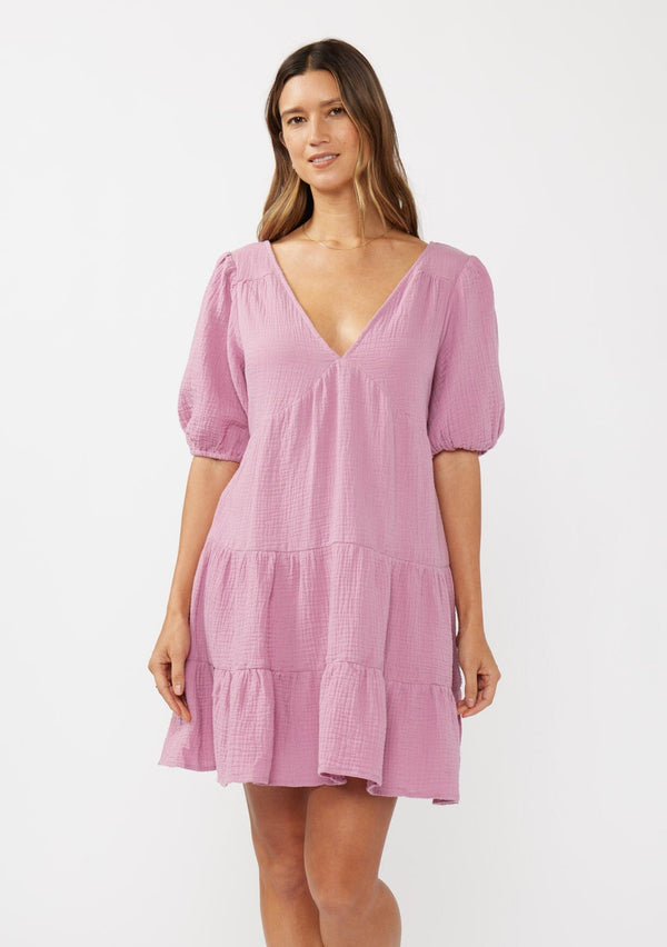 [Color: Orchid] A front facing image of a brunette model wearing a cute pink bohemian spring mini dress crafted in a soft cotton gauze. With short puff sleeves, a v neckline in the front and back, an empire waist, a tiered skirt, and an open back detail with a tie closure. 