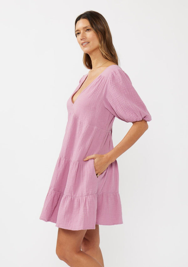 [Color: Orchid] A side facing image of a brunette model wearing a cute pink bohemian spring mini dress crafted in a soft cotton gauze. With short puff sleeves, a v neckline in the front and back, an empire waist, a tiered skirt, and an open back detail with a tie closure. 