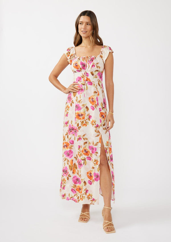 [Color: Natural/Orchid] A front facing image of a brunette model wearing a large floral print maxi dress in orange and pink florals. With a square neckline, tie front neckline detail, smocked back, flutter cap sleeves, and a side slit. A pretty spring and fall maxi dress paired with strappy sandals. 