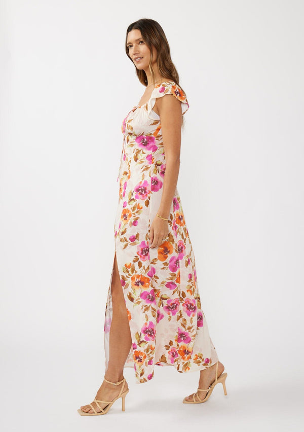 [Color: Natural/Orchid] A side facing image of a brunette model wearing a large floral print maxi dress in orange and pink florals. With a square neckline, tie front neckline detail, smocked back, flutter cap sleeves, and a side slit. A pretty spring and fall maxi dress paired with strappy sandals. 