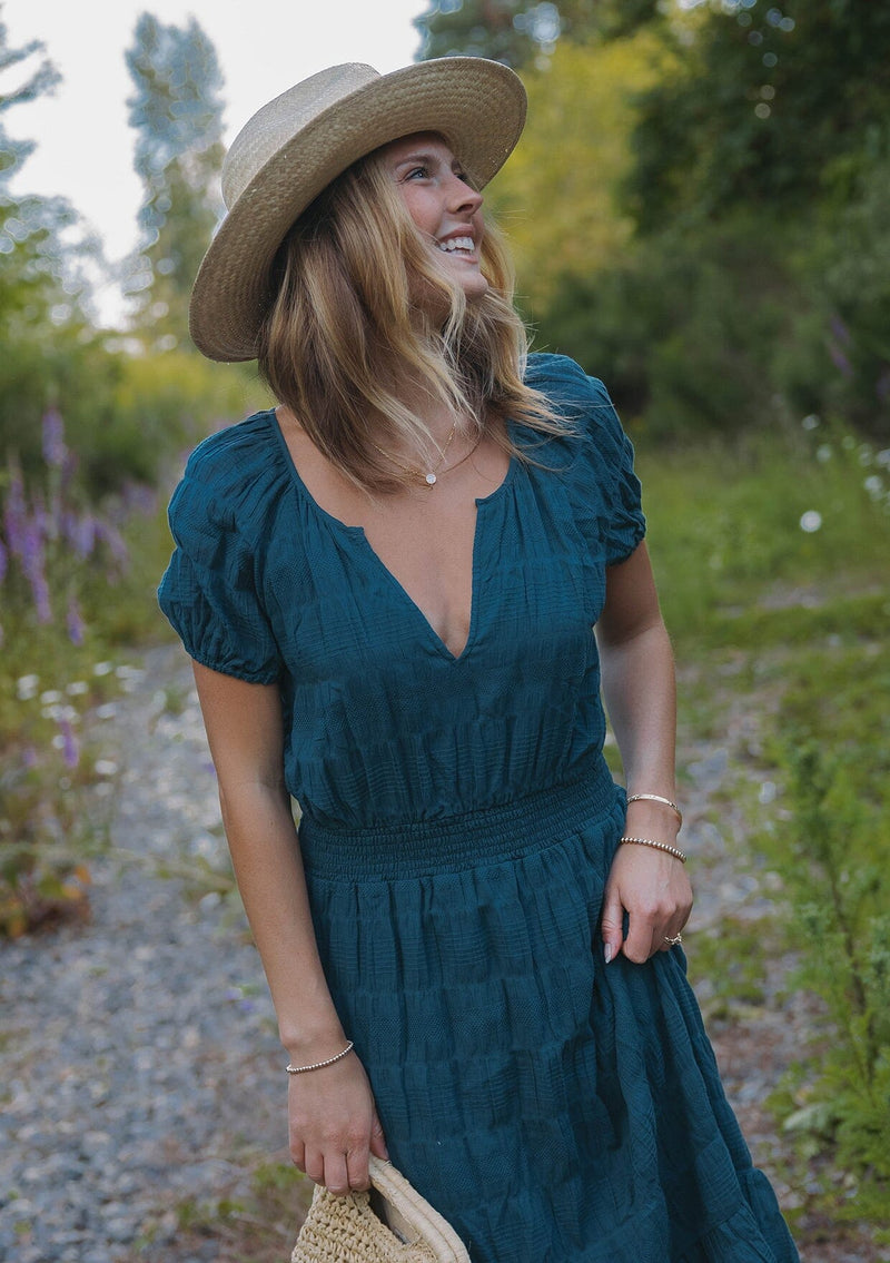 [Color: Deep Teal] A blonde model wearing a teal midi dress with a subtle woven striped design. With a split v-neckline, short puff sleeves, smocked waistline, and a ruffle trimmed hem. 