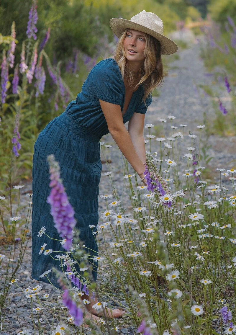 [Color: Deep Teal] A blonde model wearing a teal midi dress with a subtle woven striped design. With a split v-neckline, short puff sleeves, smocked waistline, and a ruffle trimmed hem. 