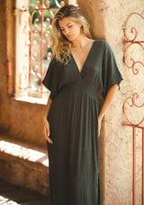 [Color: Military] An image of a blonde model wearing a classic casual military maxi dress. With half-length kimono sleeves, a plunging v neckline, a smocked elastic empire waist, side slits, and an open back with tie closure.