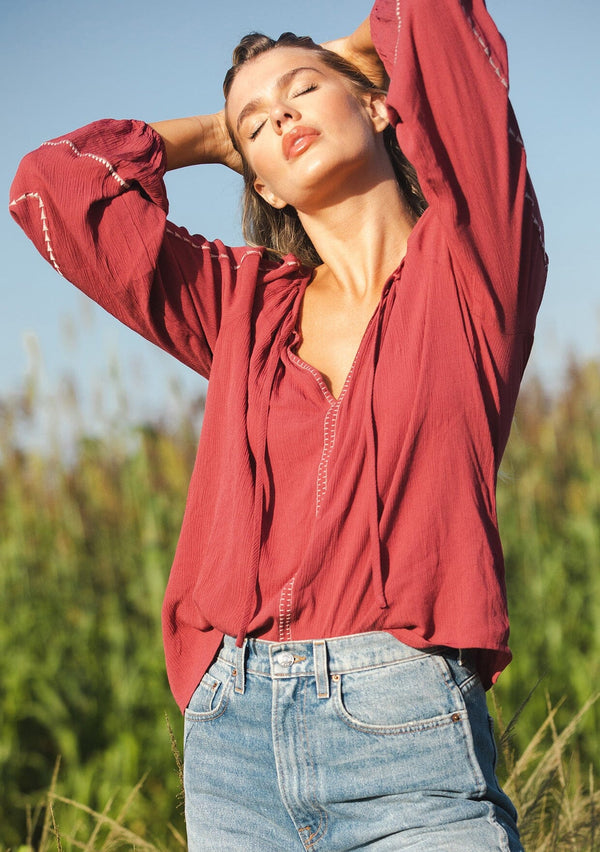 [Color: Mauve/Natural] A blonde model wearing an embroidered pink bohemian blouse. With a split v neckline with ties, long raglan sleeves, and an elastic cuff. A relaxed fit boho top styled with denim for the ultimate fall ensemble.