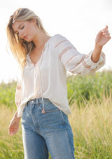 [Color: Natural/Rust] A blonde model wearing a beige ivory blouse with bohemian embroidered details. A relaxed fit blouse with a split v neckline and tassel tie front, long sleeves, and ruffle trim details at the neck. Styled with denim for an ultra boho look for the fall season.