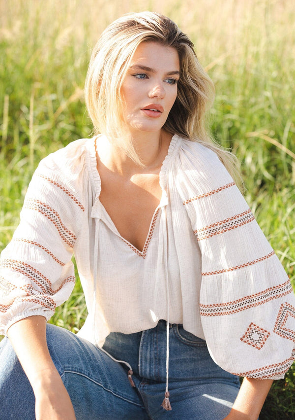 [Color: Natural/Rust] A blonde model wearing a beige ivory blouse with bohemian embroidered details. A relaxed fit blouse with a split v neckline and tassel tie front, long sleeves, and ruffle trim details at the neck. Styled with denim for an ultra boho look for the fall season.