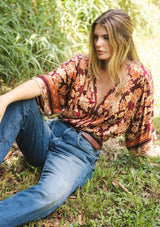 [Color: Wine/Dusty Rose] A blonde model wearing a orange and red bohemian floral border print blouse. Perfectly suited for the fall season, with flowy kimono sleeves, button front, flattering v neckline, and half smocked elastic waist at the back. A trendy and stylish boho top paired with vintage denim. 