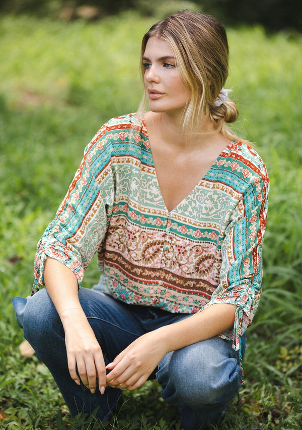 [Color: Jade/Natural] A blonde women wearing a multi print bohemian top with a v neckline, button front, ruched long sleeves, and elastic hemline. A teal and green causal summer top styled with denim for a summer to fall transition.