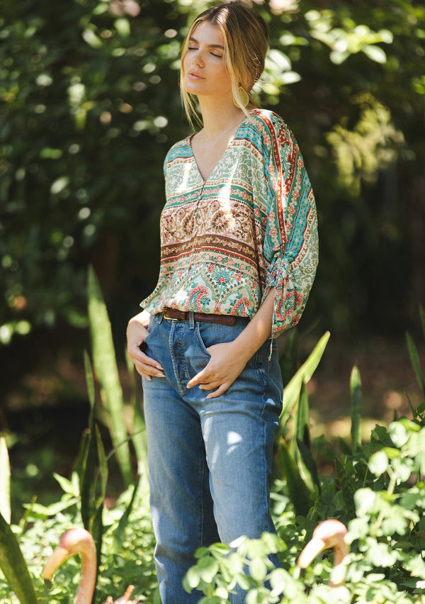 [Color: Jade/Natural] A blonde women wearing a multi print bohemian top with a v neckline, button front, ruched long sleeves, and elastic hemline. A teal and green causal summer top styled with denim for a summer to fall transition.