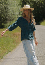 [Color: Deep Teal] A blonde model wearing a textured jacquard stripe blouse in teal. A relaxed fit blouse with a split v neckline and long sleeves. Styled with loose fit denim jeans.