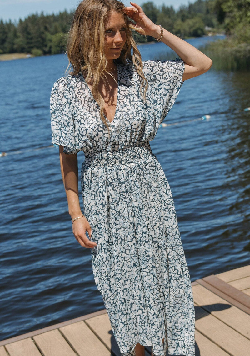 [Color: Teal/Taupe] A blonde woman wearing a summer to fall transitional midi dress with floral print on a chiffon like fabric. With a flattering v neckline, smocked waistline and an asymmetrical hem with multiple slits.