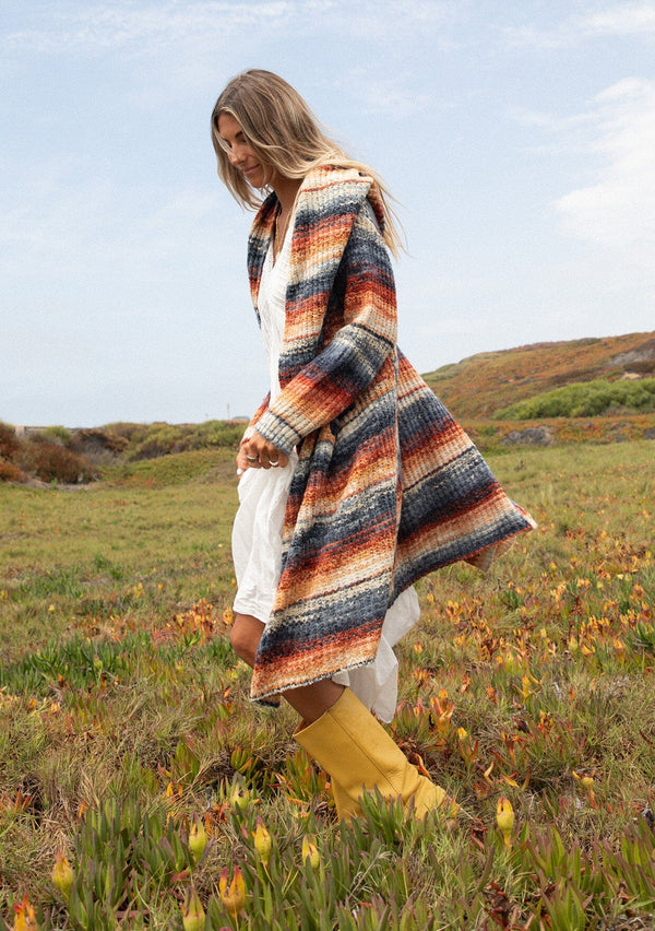 [Color: Rust/Navy] A side facing image with a blonde model wearing a cozy bohemian space dye chunky knit cardigan in a rust multi-color stripe pattern. This plush cardigan features long sleeves, a cozy hood, essential pockets, and an open front. The long length and thick knit make it the perfect sweater for the fall season.