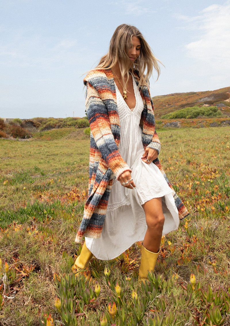 [Color: Rust/Navy] A front facing image with a blonde model wearing a cozy bohemian space dye chunky knit cardigan in a rust multi-color stripe pattern. This plush cardigan features long sleeves, a cozy hood, essential pockets, and an open front. The long length and thick knit make it the perfect sweater for the fall season.