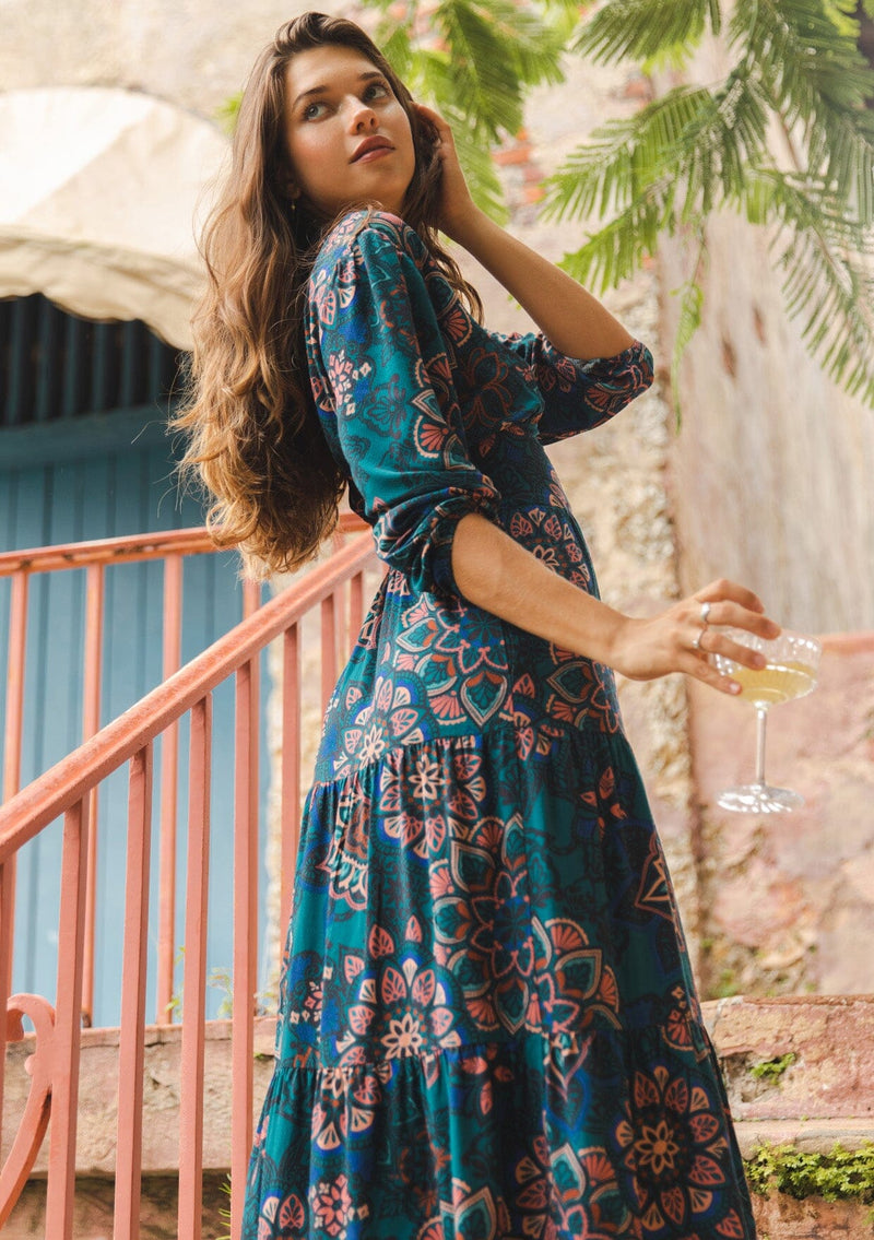 [Color: Teal/Rust] A brunette model wearing a tiered midi dress in a teal floral print. With a slim fit bodice, v neckline, long sleeves, and convenient side pockets. The sexy open back with tie detail completes this everyday dress. Paired with flats for a relaxed style, perfect for the Fall season.